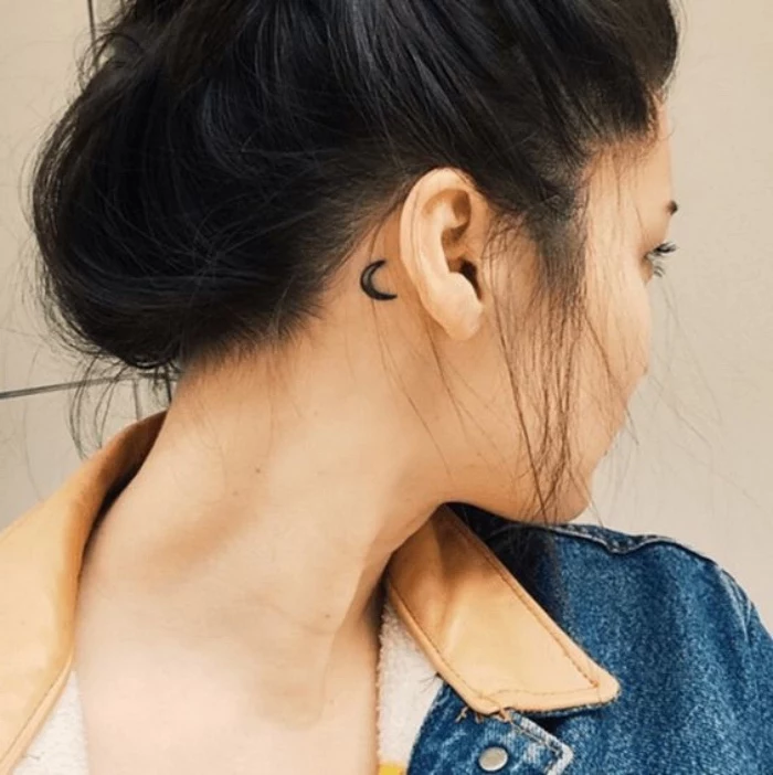 crescent moon behind the ear tattoo, small tattoos for women, black hair in a messy bun, jean jacket