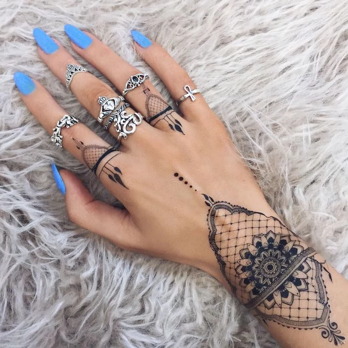 long blue nail polish, mandala tattoos, finger tattoo ideas, many silver rings, hand resting on a furry grey blanket