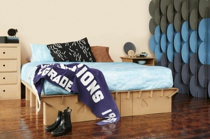 cardboard bed, with blue linens, on a wooden floor, cardboard dresser, cardboard night stand