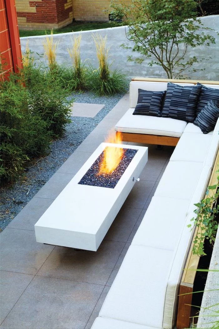 cement table, with a small fire pit, garden decoration ideas, corner sofa, with black throw pillows, planted bushes
