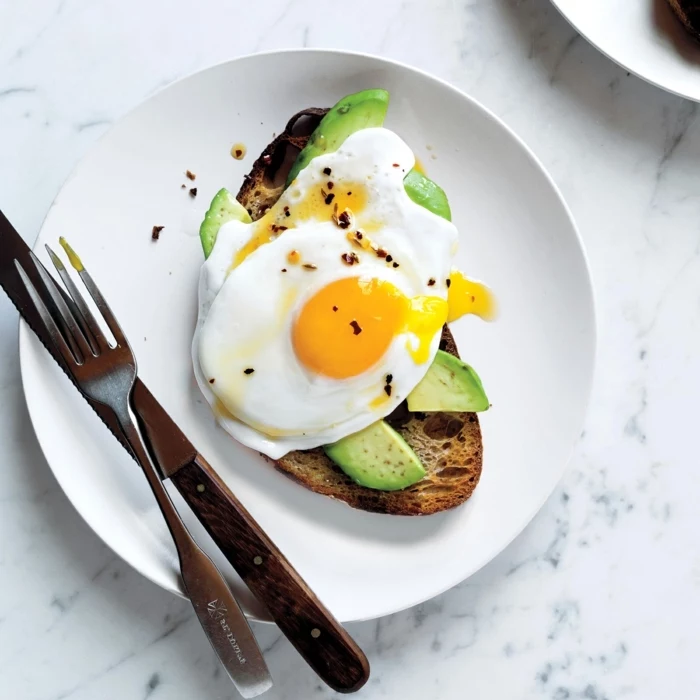 toasted bread, with avocado slices, eggs on top, balanced meal, on a white plate, knife and fork on the side
