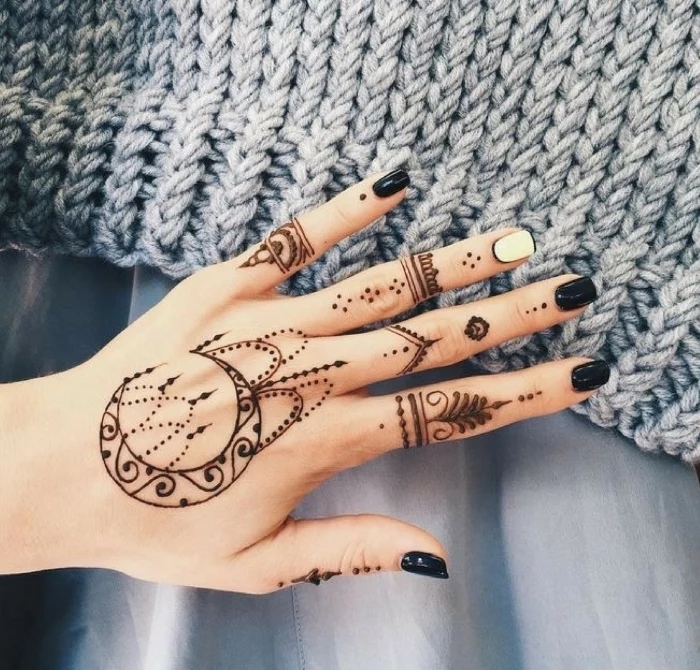 large henna tattoo, fingers crossed tattoo, hand resting on a grey blanket, with black nail polish