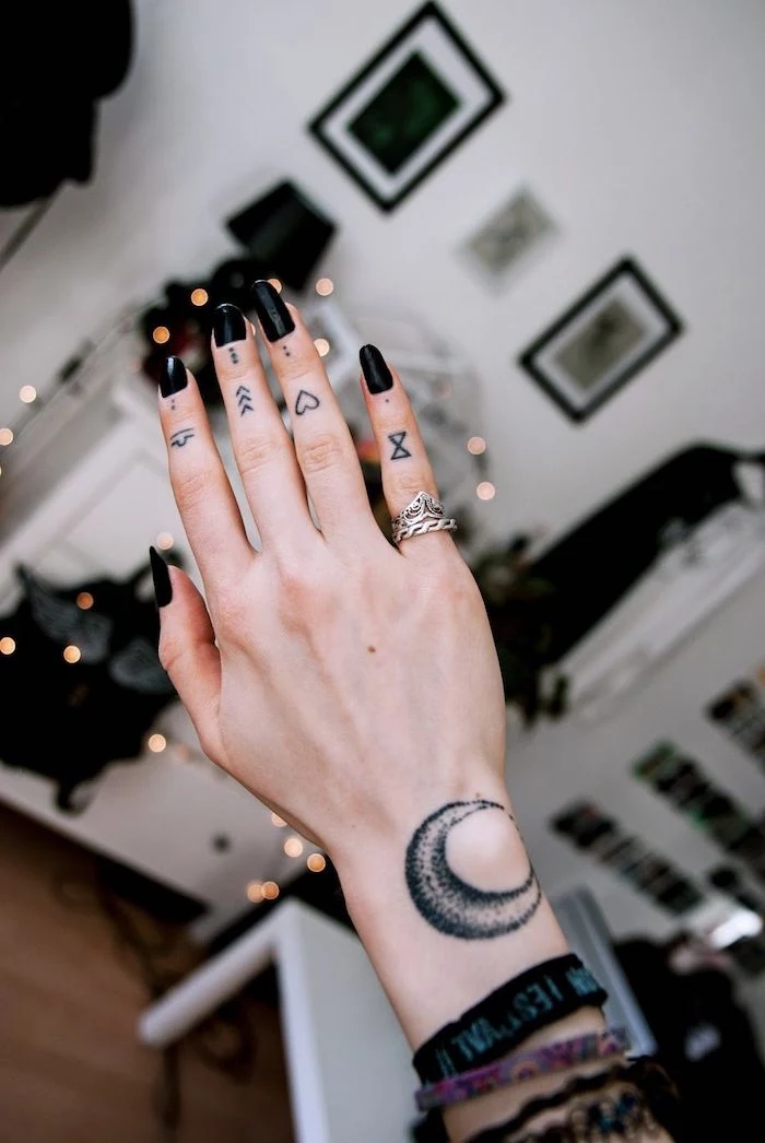 hand with different tattoos on each finger, black nail polish, finger tattoo, picture frames in the background