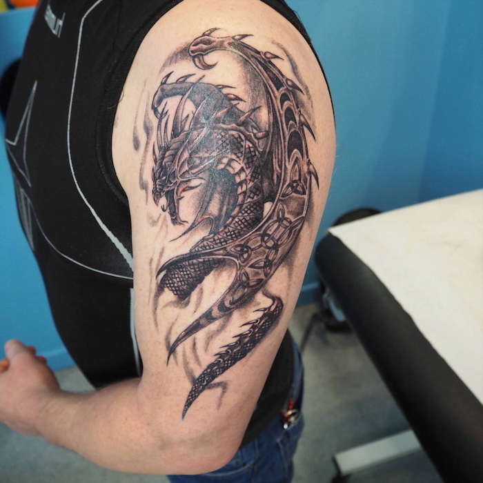 large dragon, shoulder tattoo, wrist tattoos for men, man wearing a black top and jeans, blue background