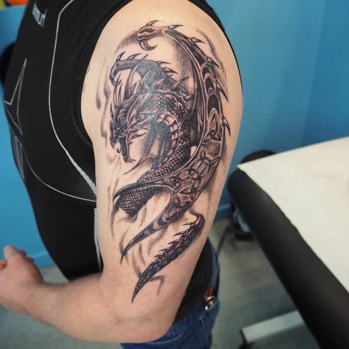 large dragon, shoulder tattoo, wrist tattoos for men, man wearing a black top and jeans, blue background