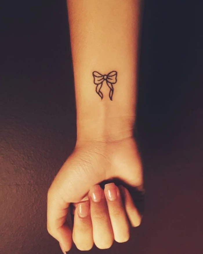 small bow wrist tattoo, small tattoo ideas for men, hand clutched in a fist, in front of a black background