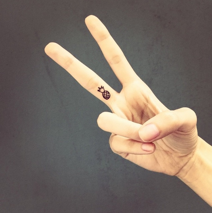 small pineapple finger tattoo, person making a peace sign, small butterfly tattoos, in front of a grey background