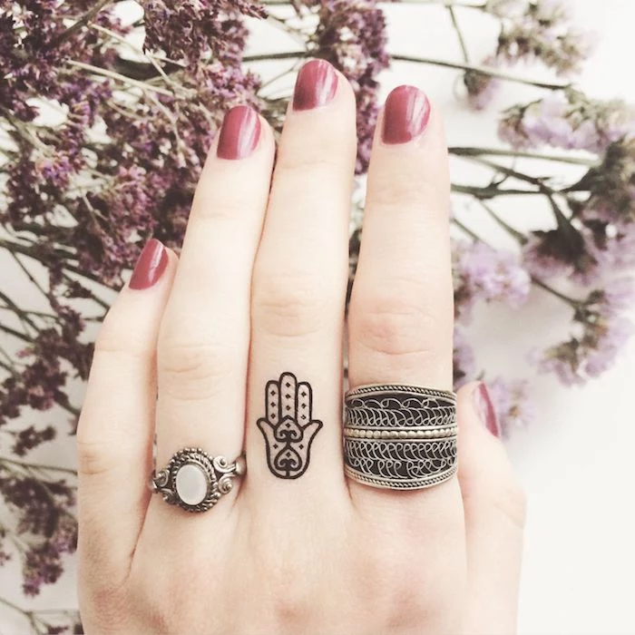 hand tattoo, middle finger tattoo, silver rings, red nail polish, lion finger tattoo, flowers in the background
