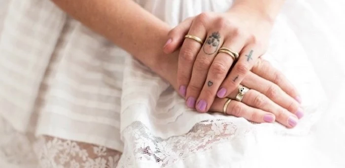 hands resting on a white dress, cross heart and skull, finger tattoos, rose finger tattoo, pink nail polish