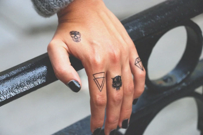 hand resting on a metal railing, lots of different tattoos on the fingers, black nail polish, finger tattoos