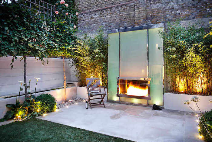large fireplace, a small chair next to it, on a cement floor, small backyard landscaping, planted trees bushes and flowers