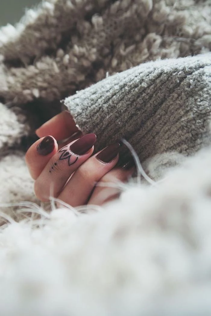lotus flower middle finger tattoo, finger tattoo, burgundy nail polish, hand resting on a grey blanket