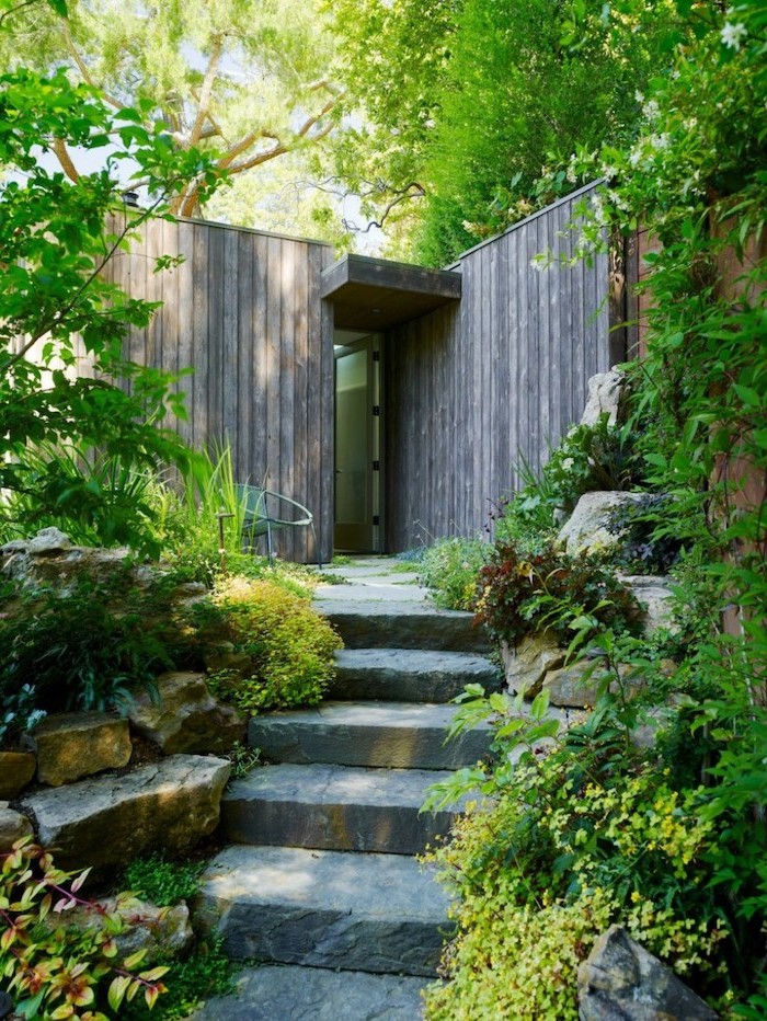 stone pathway, planted bushes flowers and trees alongside it, small yard landscaping, wooden entrance