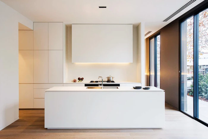 white minimalistic kitchen, white kitchen island in the middle, kitchen appliances, white cupboards and drawers, large windows