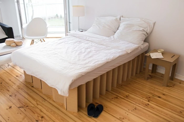cardboard bed, with white linens, on a wooden floor, with a cardboard nightstand, cardboard chair