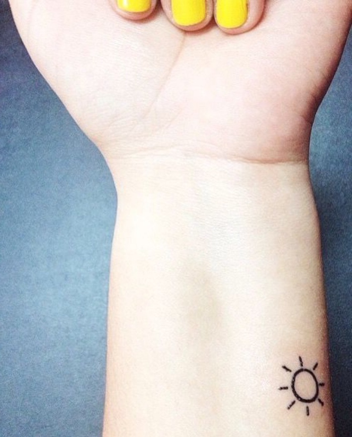 small sun wrist tattoo, best small tattoos for men, woman with yellow nail polish, in front of a blue background