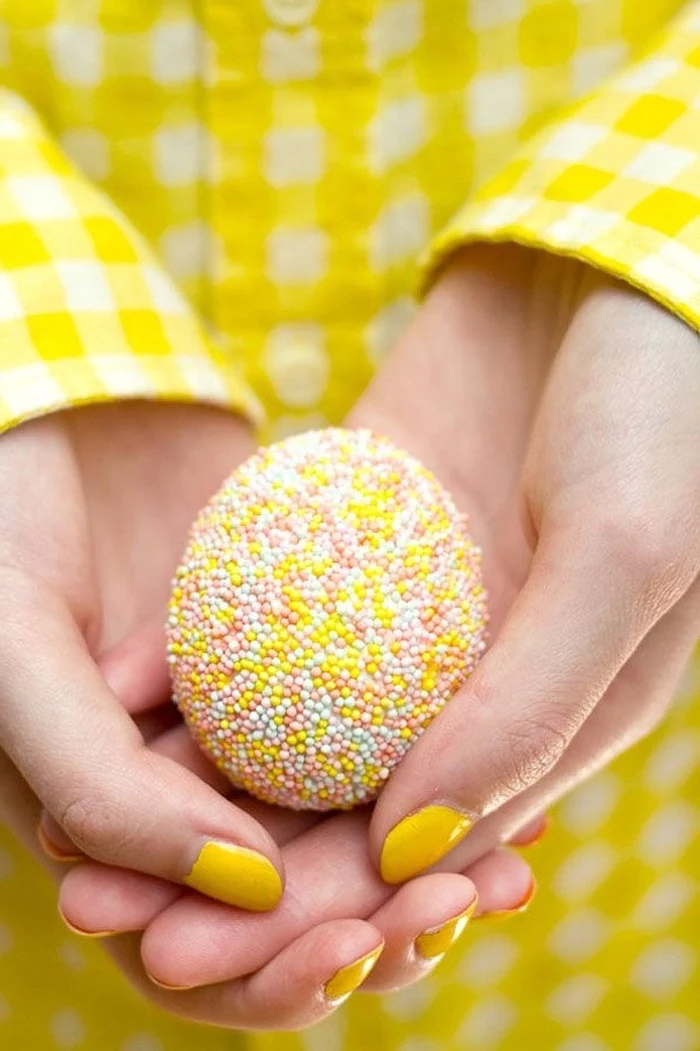 hand with yellow nail polish, wearing a yellow plaid shirt, holding a sprinkled egg, how to color easter eggs