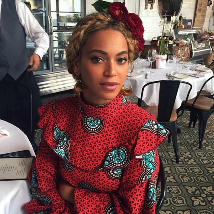 beyonce sitting down, wearing a glower wreath headband, african dresses, patterned top, braided hair