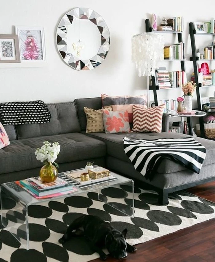 light gray walls, grey corner sofa, colourful printed, throw pillows, wooden bookshelves, glass coffee table