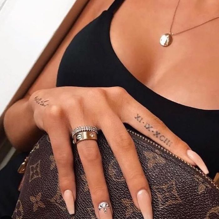 black top, finger tattoo, long pink nails, roman numerals tattoo on chest, gold necklace and rings