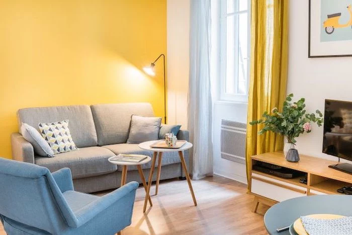 yellow accent wall, grey sofa, how to decorate a small living room, blue armchair, small wooden coffee tables