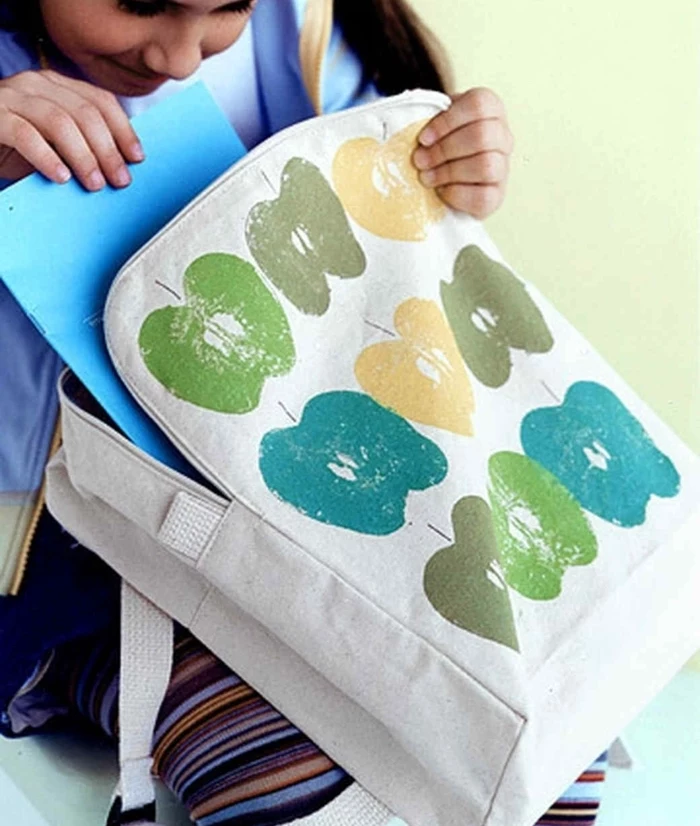white backpack, apple prints on it, green blue and yellow, prek learning games, little girl holding it