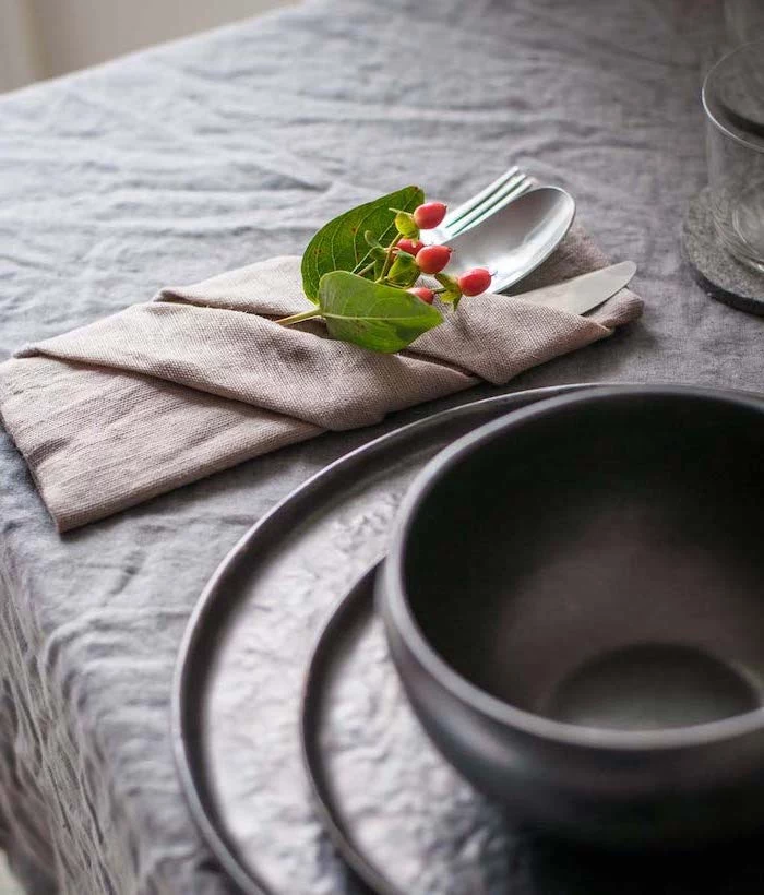 grey table cloth, beige folded napkin, silverware inside, thanksgiving napkin folding, black bowl and plates