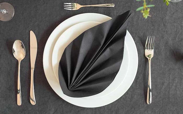 black napkin, in the shape of a leaf, how to fold napkins, silverware arranged, around white plates