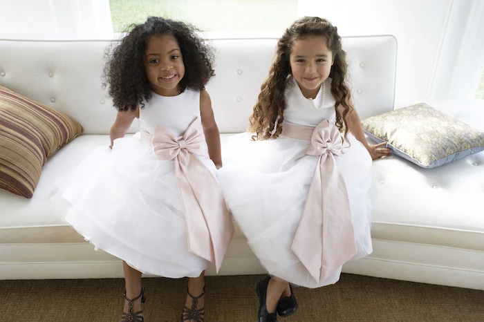 two girls sitting on a white sofa, wearing white dresses, with ivory bows, flower girl hair, curly hair