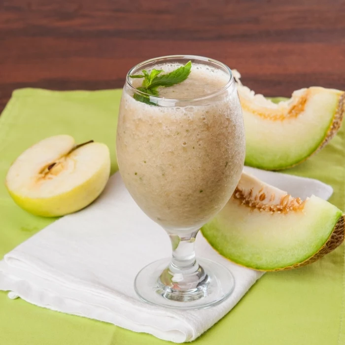 smoothie recipes with frozen fruit, sliced melon and apple, tall glass, on a white cloth, mint on top