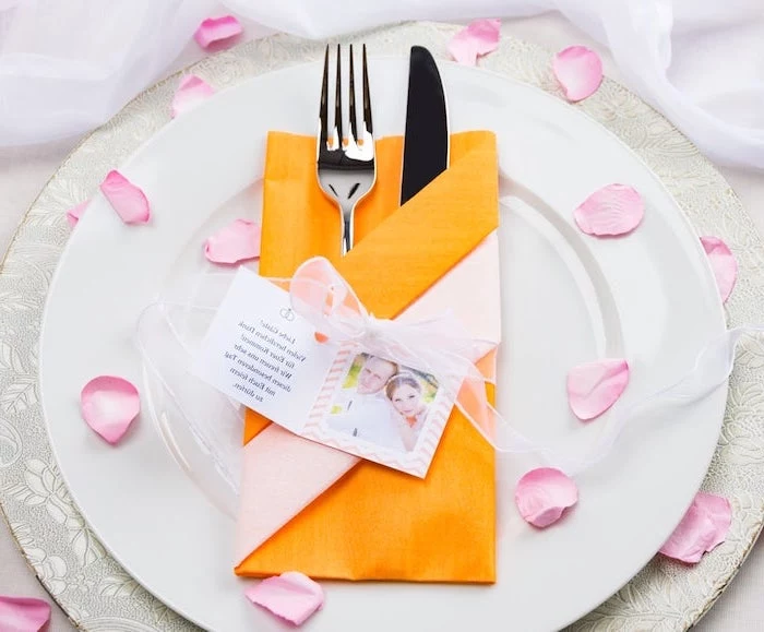 how to fold cloth napkins, orange napkin, silverware inside, on white plates, rose petals scattered around