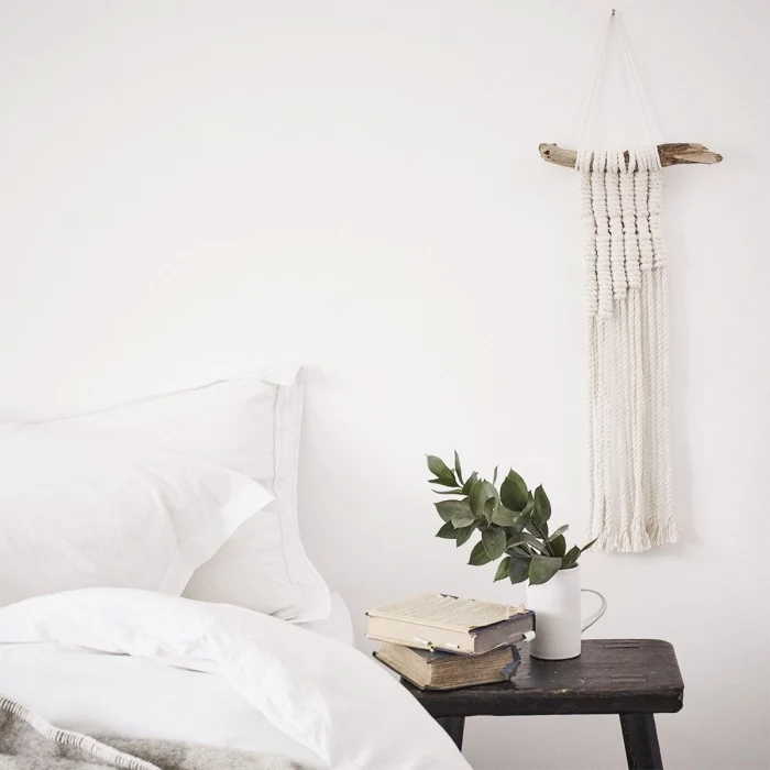 wooden night table, two books, ceramic pot, white bed linen, macrame tapestry