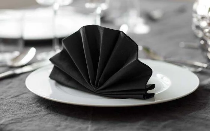 fan shaped, black napkin, napkin folding with silverware, on a white plate, black table cloth