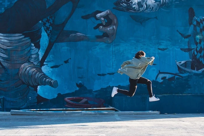 wall covered in blue graffiti, woman running, wearing white sneakers black pants, grey jacket