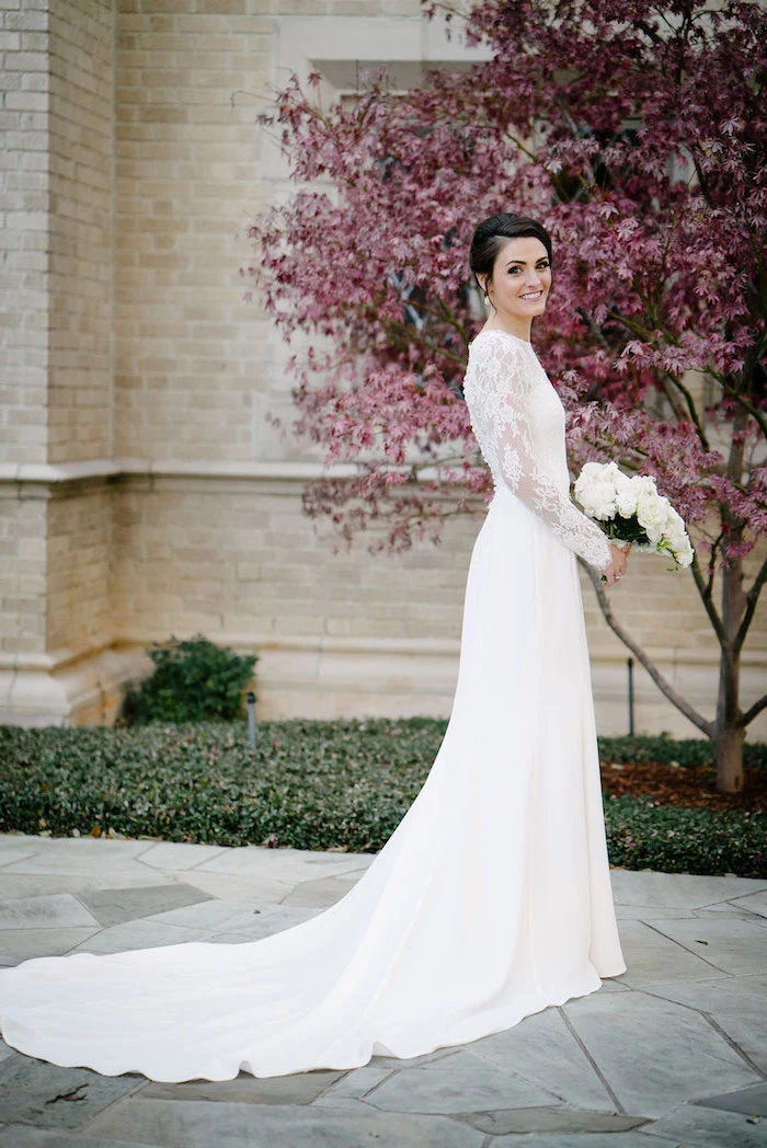 a line wedding dresses, white lace dress, long train, black hair, in a low updo, pink blooming tree, white flowers bouquet