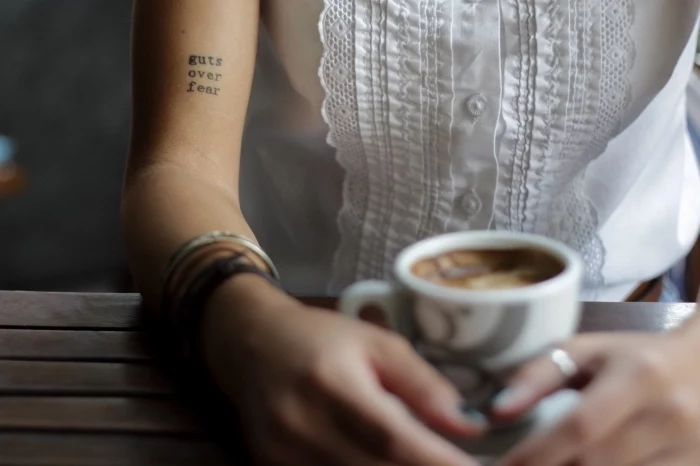 guts over fear, coffee cup, white shirt, inside arm tattoo, tattoo locations, wooden table