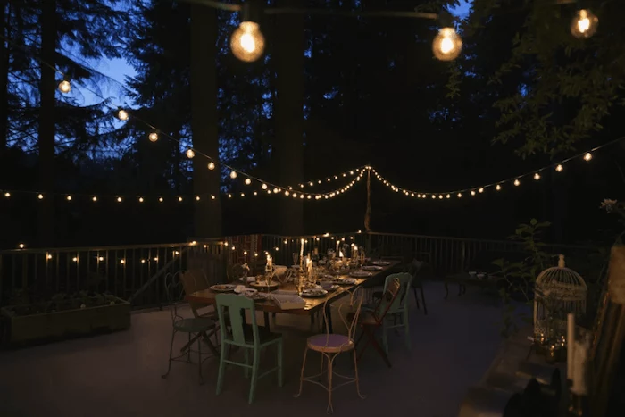 strings of lights, metal chairs, covered porch ideas, wooden table, metal railing, tall trees