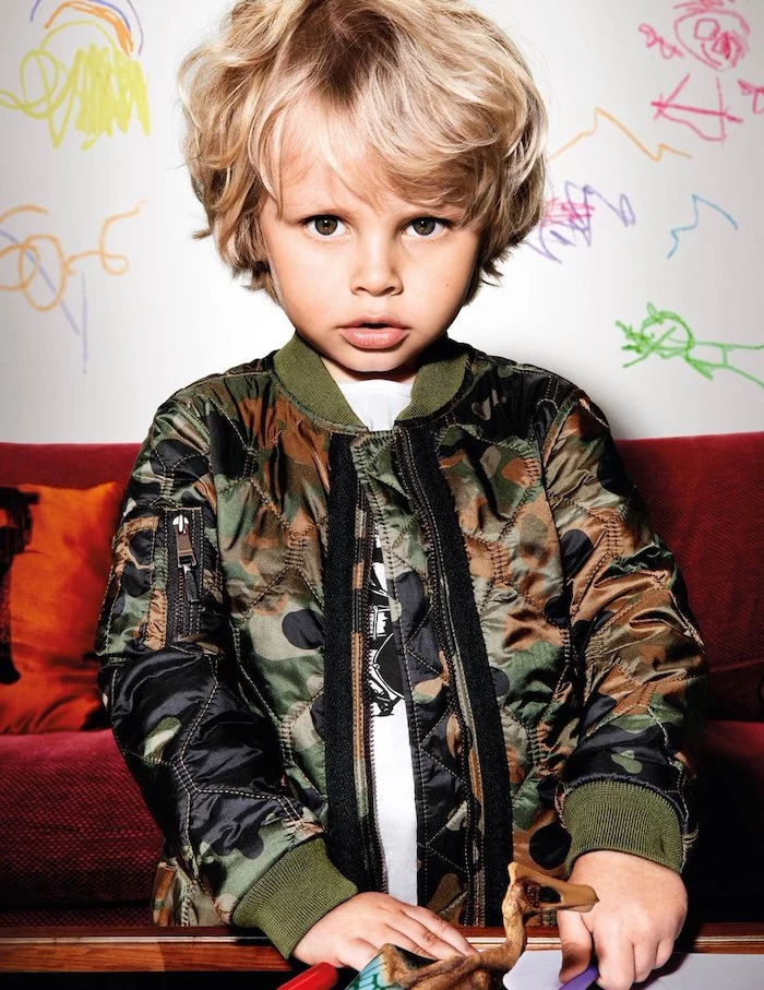 little boy, with blonde hair, navy jacket, teen boy haircuts, red sofa, white wall, with drawings