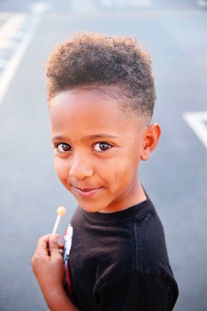 little boy smiling, holding a lollipop, wearing a black shirt, teen boy haircuts, black curly hair