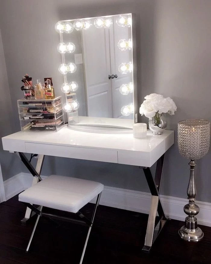 small makeup vanity, white table, white leather stool, mirror with lights, grey wall, acrylic makeup drawers