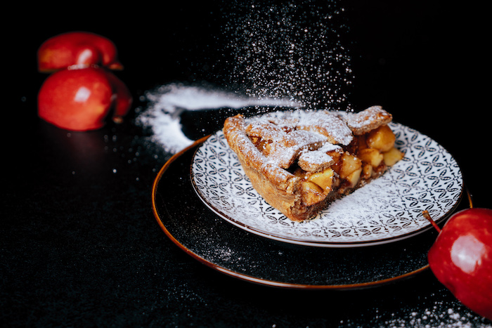 apple pie slice, dusted with powdered sugar, placed on black and white plate, apple pie recipe, halved apples on the side