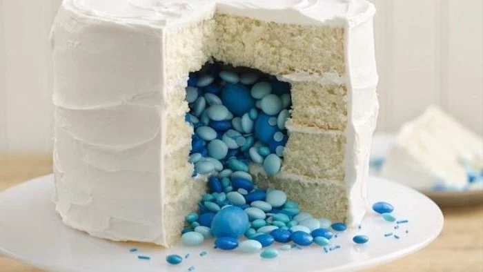 small cake, with white frosting, blue candy inside, gender reveal balloons, white cake stand