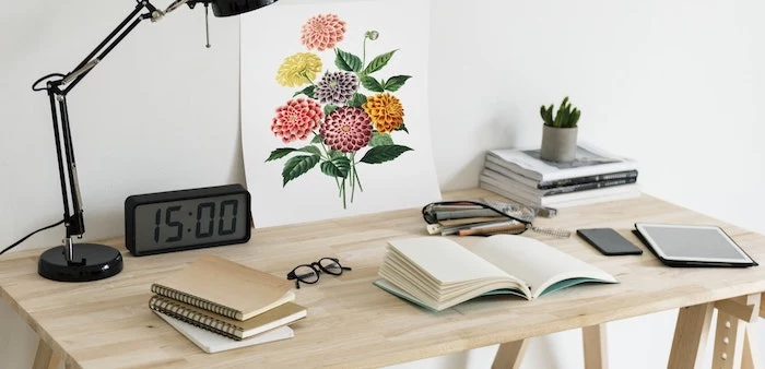 wooden desk, covered in books, digital clock, floral art, cubicle accessories, black lamp, white wall