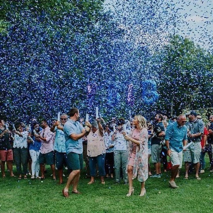 gender reveal decorations, lots of people smiling, standing on a field, blue confetti in the air