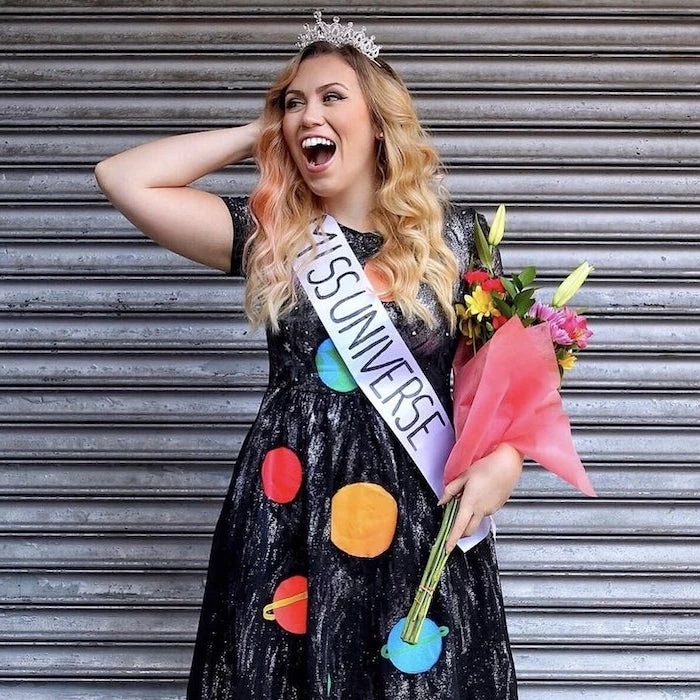 woman smiling, halloween costumes ideas for adults, wearing a black dress, miss universe sash, flowers and tiara