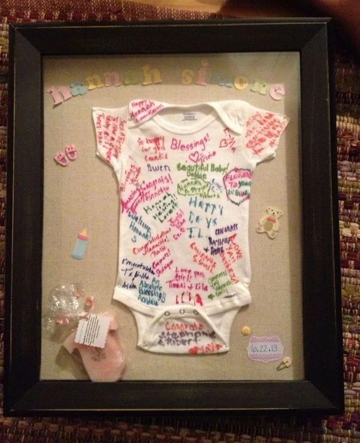 white onesie, with different things written on it, framed in black frame, hannah simone, baby shower table decorations
