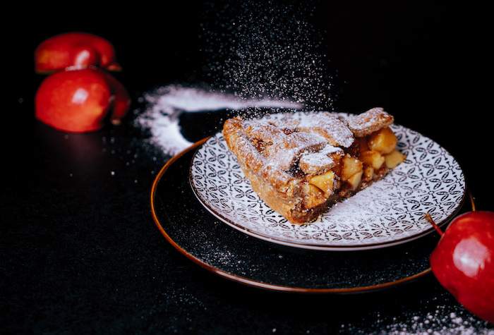 apple pie recipe, slice of apple pie, dusted with powdered sugar, placed on black and white plate, halved apples on the side