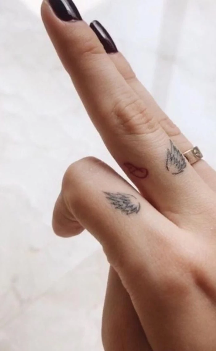 black nail polish, white background, gold ring, finger tattoos, wing tattoo on arm