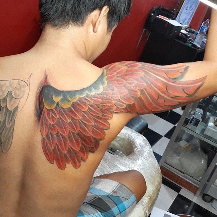 man with black hair, sitting on a bed, wings neck tattoo, red wing, back tattoo, extending to the arm