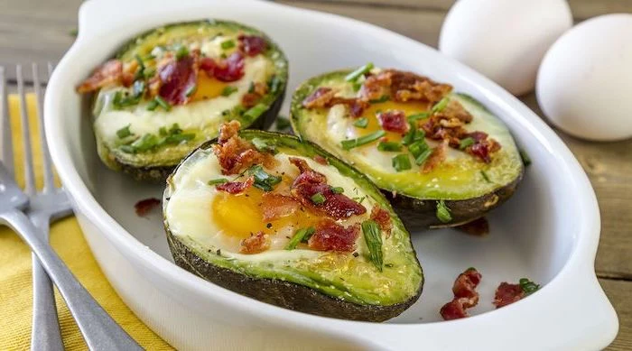 halved avocados, filled with eggs, chives and bacon on top, white casserole, low carb breakfast ideas, yellow table cloth
